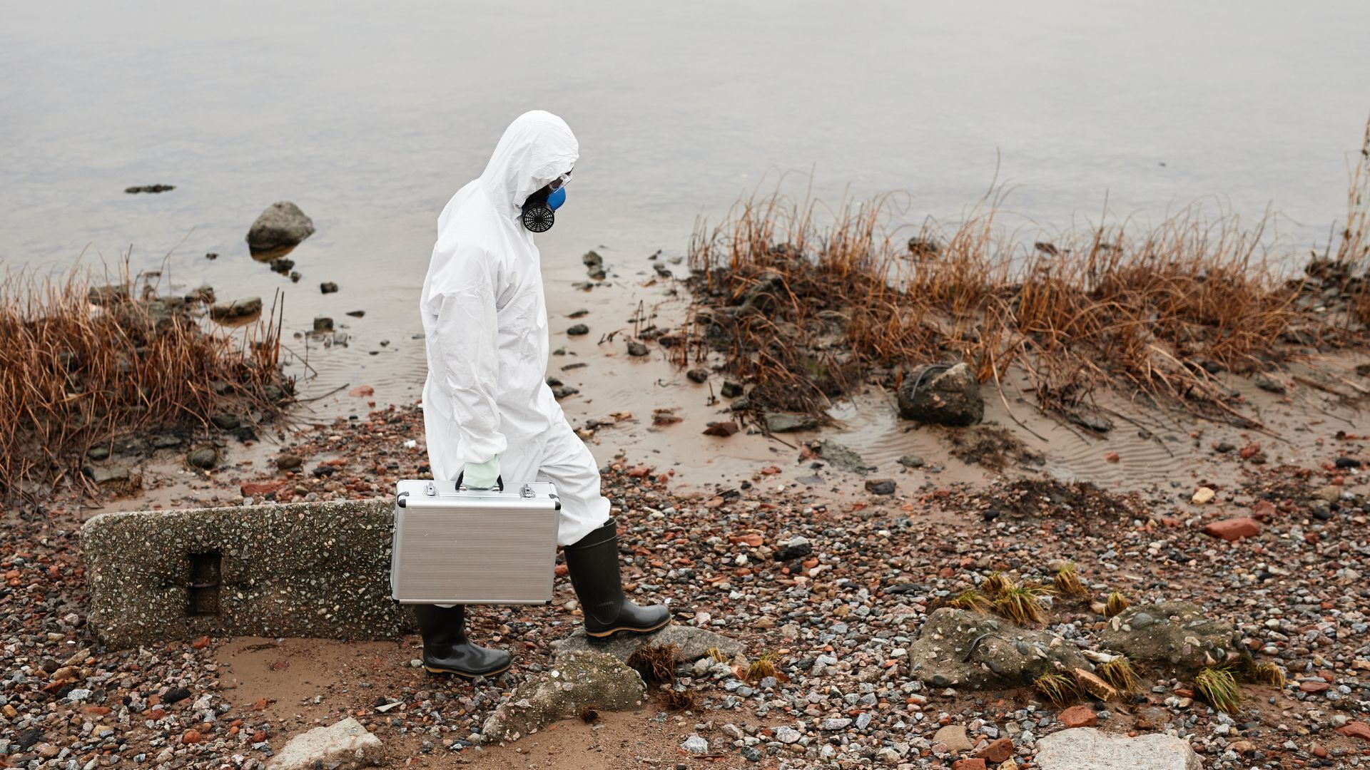 person in white coveralls with boots and briefcase walking along riverbank