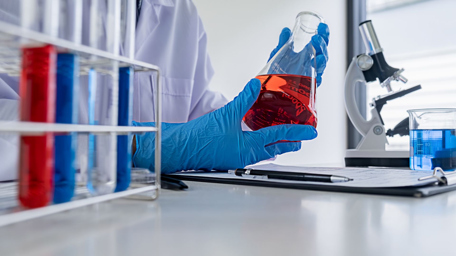 scientist in lab coat with blue gloves holding vial with red liquid